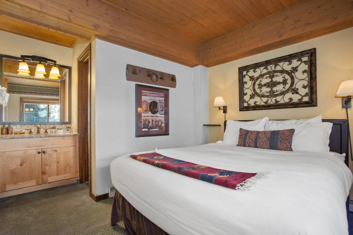 A cozy master bedroom with a queen-sized bed dressed in soft bedding, decorative wall art, and a vanity area with a mirror. Warm sunlight streams in, enhancing the inviting and intimate feel of the space—perfect for reading or morning coffee.