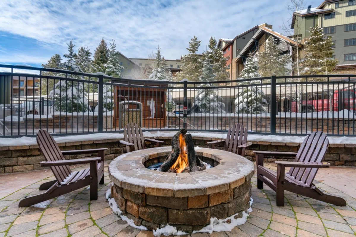 A snow-covered outdoor fire pit area with chairs arranged for socializing, set among wintry trees. The inviting glow of the fire contrasts with the surrounding snow, creating a perfect spot for sharing stories and warming up.