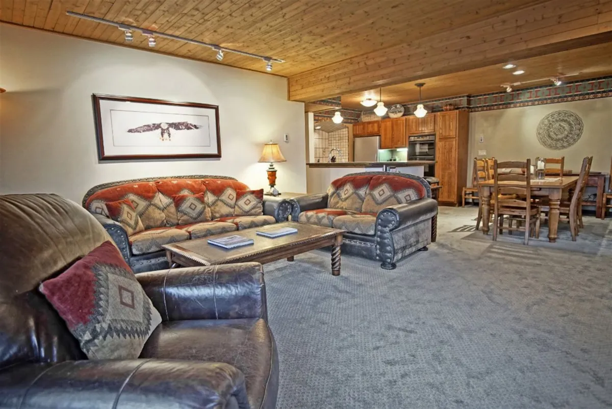 This image features a warm, open-concept living area with plush leather sofas, a coffee table with books, a visible dining area in the background, framed artwork, and soft lighting from a lamp. The arrangement showcases seamless flow between living and dining zones, ideal for relaxation and socializing.