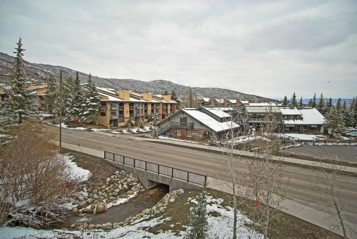 An inviting winter exterior scene featuring snow-covered mountains in the background, a wooden bridge over a creek, and nearby residential buildings. The serene mood is complemented by visible ski tracks and proximity to the base area, evoking the ski-in/ski-out lifestyle.