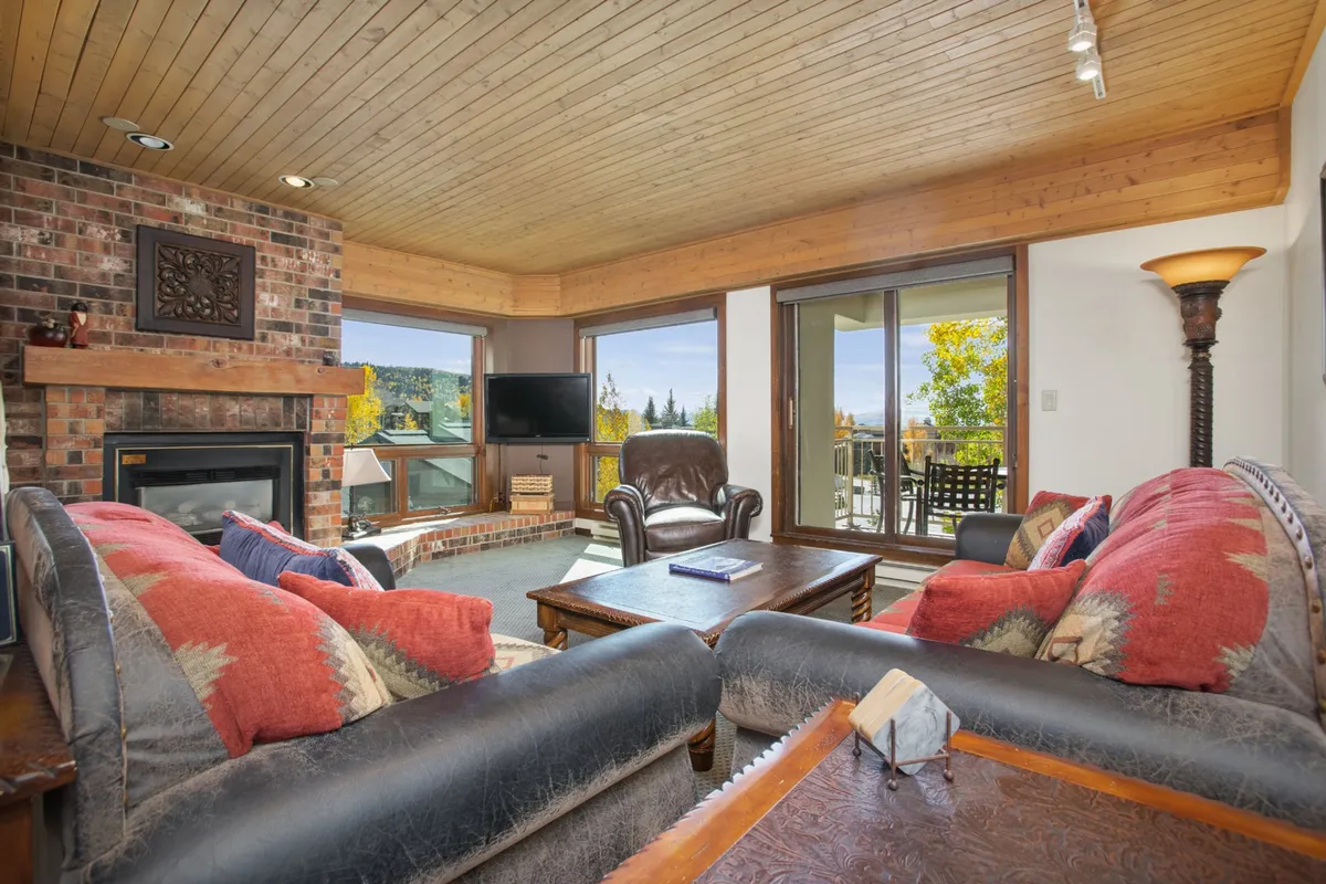 A cozy living room scene with a prominent fireplace, a sofa with pillows, and large windows offering a glimpse of the outdoors. The fire is lit, and the space exudes warmth and comfort, perfect for relaxing after a day on the slopes.