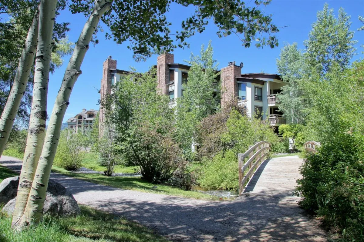 This image captures a peaceful exterior scene featuring a charming wooden bridge surrounded by lush greenery and mature aspen trees, with the Dulany building facade in the background. The area evokes a sense of natural adventure and relaxation, suggesting easy access to walking paths, creekside fun, and outdoor activities.
