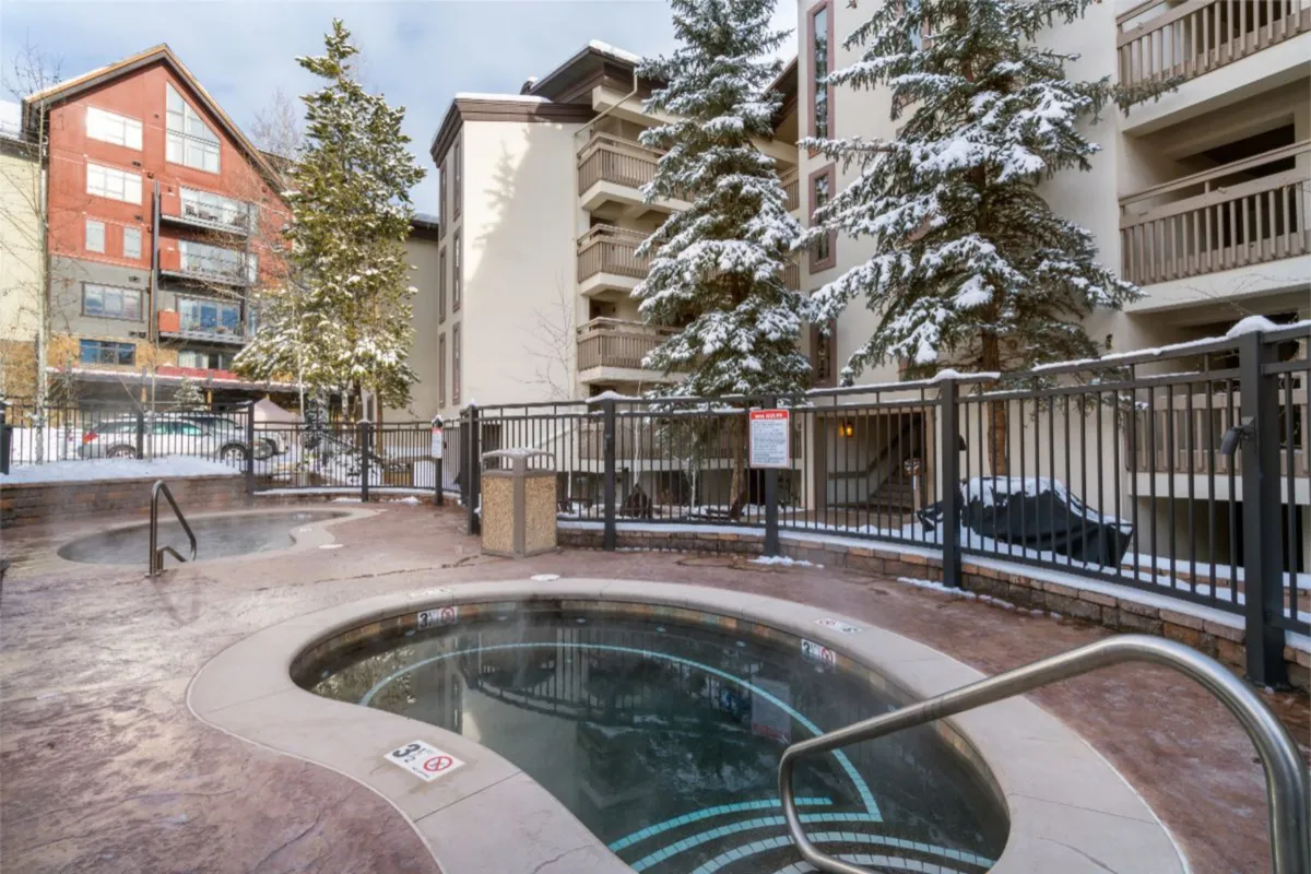 The outdoor hot tub area in winter, surrounded by snow-laden trees and the Dulany building. The scene evokes relaxation and camaraderie after a day on the slopes, with steam rising from the hot tub and a cozy mountain lodge feel.