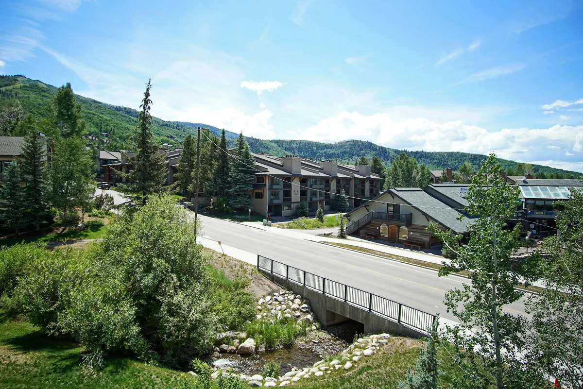 A lush, green summer landscape with mountains in the distance, tree-lined pathways, and nearby community buildings. The scene suggests hiking, biking, and community events, inviting exploration of nature and local festivities in warmer months.