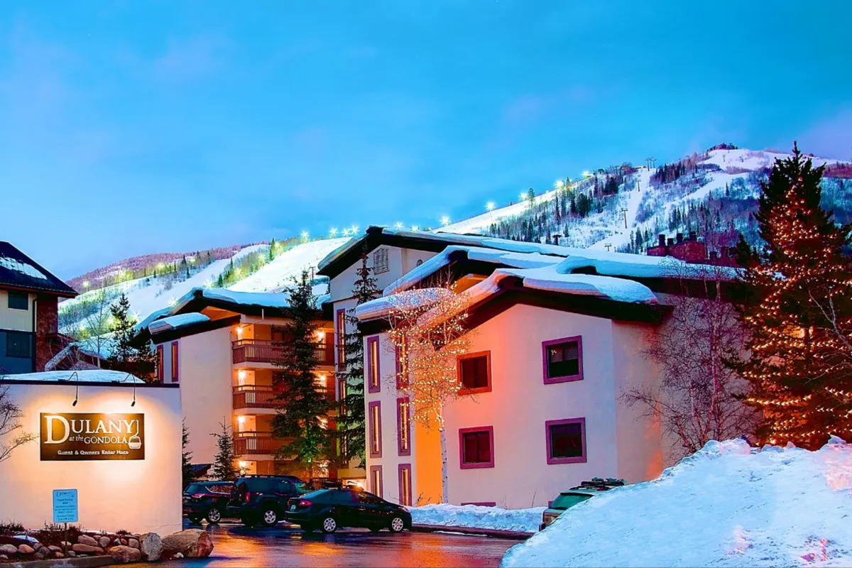 A festive winter exterior of the Dulany building with twinkling lights adorning snow-covered trees, the Dulany sign visible in front, and a majestic snow-capped mountain as a backdrop. The entry area is surrounded by fresh snow, evoking a classic ski resort ambiance.