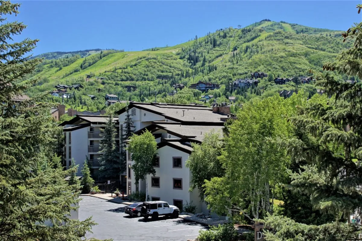 The image features a dramatic view of the mountains, trees, and the building’s architecture, highlighting the property’s proximity to outdoor adventure. The scene emphasizes expansive mountain vistas and a tranquil, nature-rich environment.