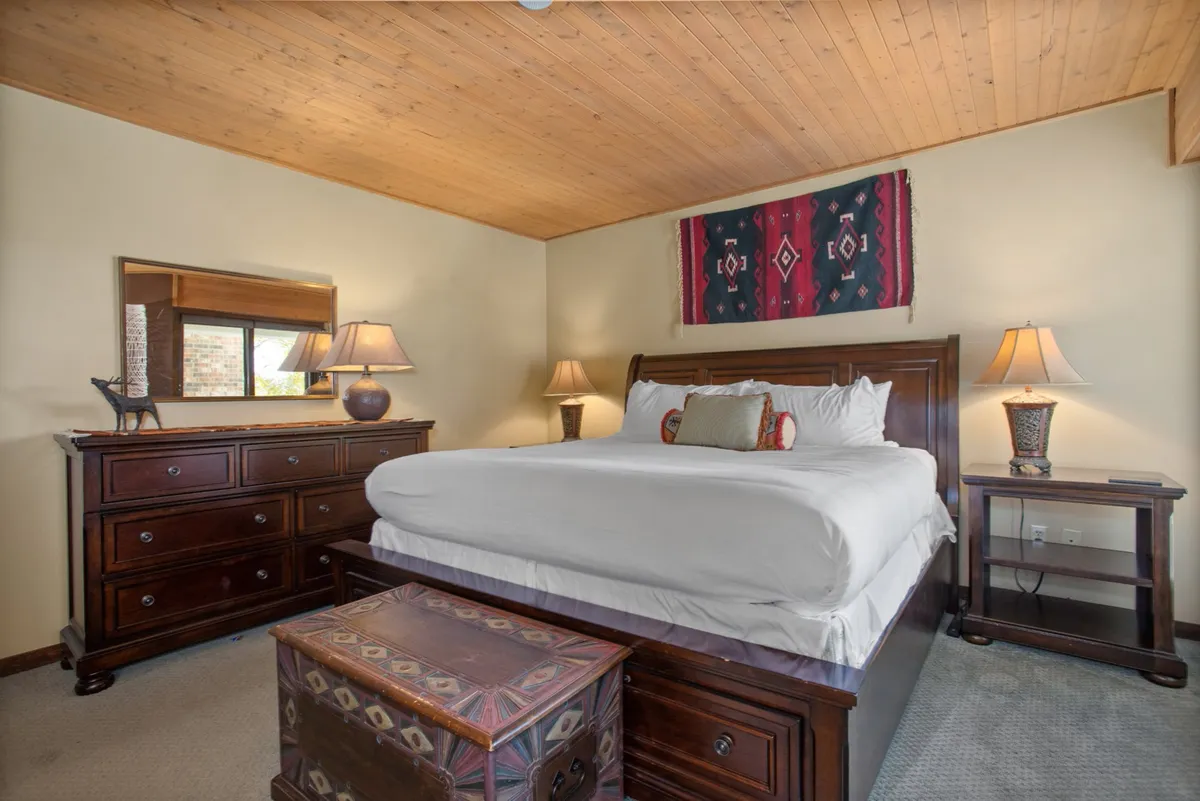 A tranquil, restful master suite featuring a king-sized bed with plush bedding, decorative chest, bedside lamps for reading, and a woven wall hanging. Soft morning light fills the room, creating an inviting and private atmosphere.