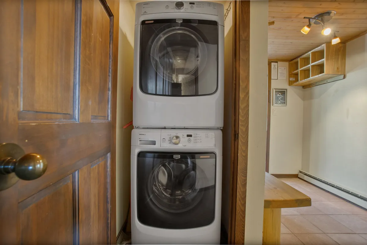 The interior scene focuses on a stacked washer and dryer within a clean, organized laundry nook, complete with wooden shelves for storage and a sturdy wooden door. The space is functional yet inviting, highlighting the convenience of in-unit laundry.