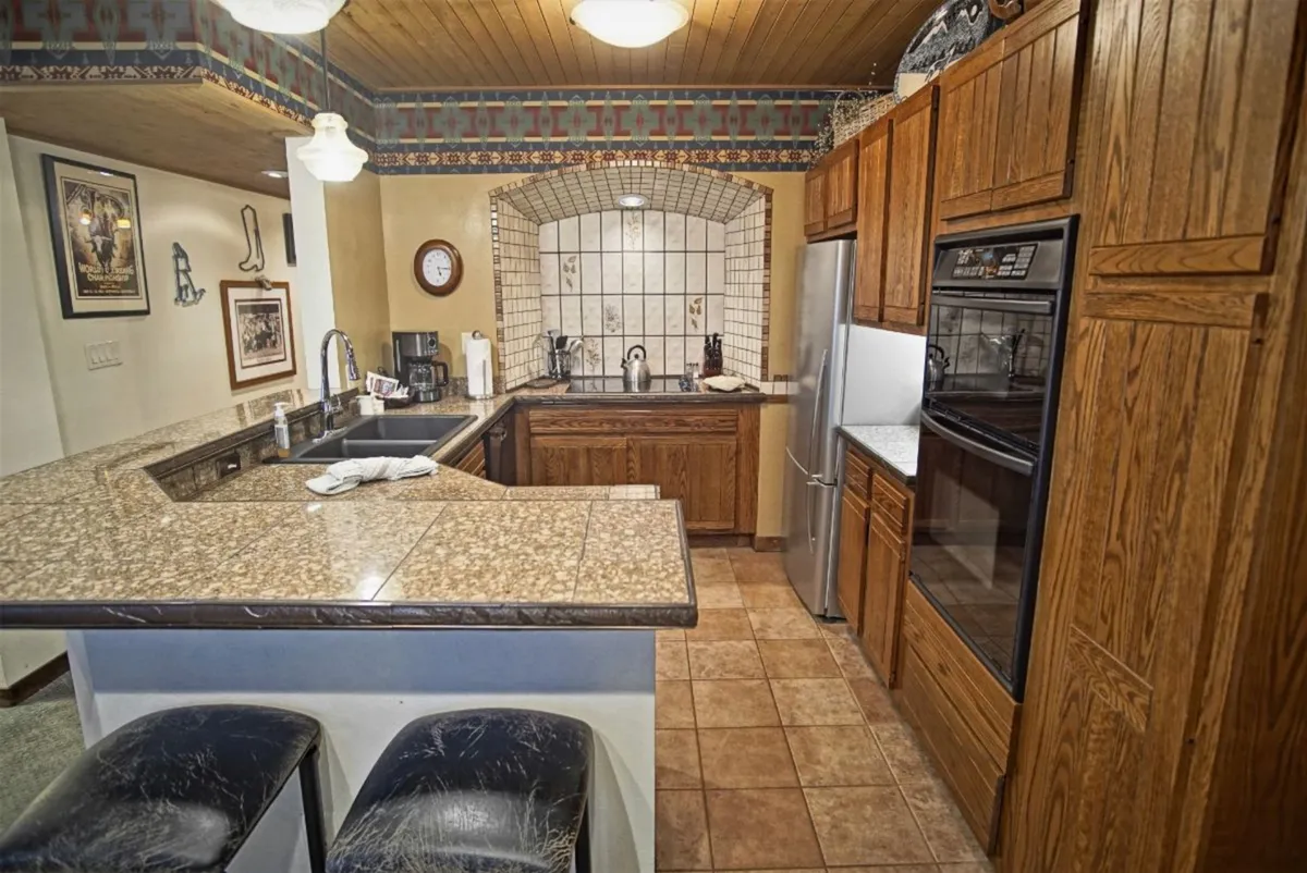 A modern, fully equipped kitchen with granite countertops, wood cabinetry, decorative tile backsplash, stainless steel appliances, and bar stools. The kitchen is clearly designed for group meal preparation and casual conversation.