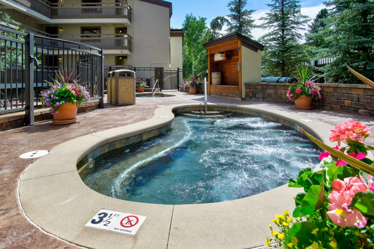 An inviting outdoor hot tub is featured front and center, bordered by flower pots and lush greenery. The open setting encourages relaxation and social interaction, with enough room for friends or family to enjoy the space together.
