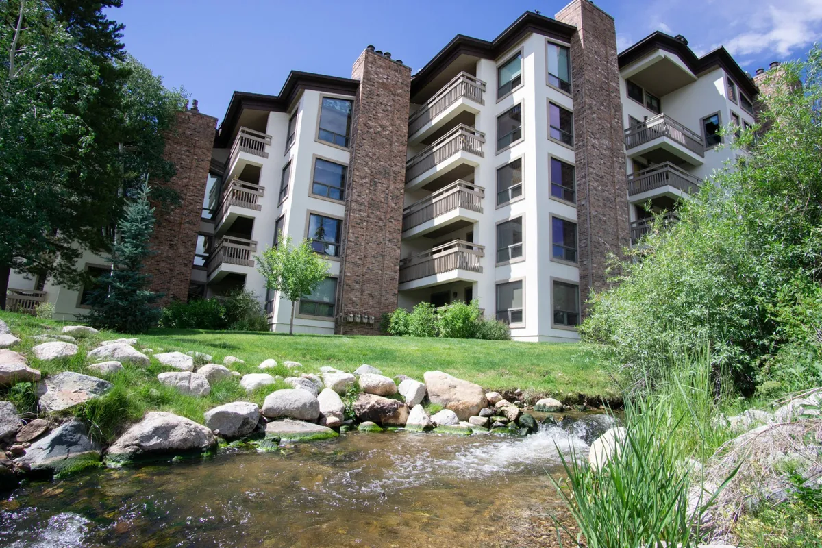 The image shows the exterior of a stylish condo building nestled beside a gentle stream, framed by lush greenery and mature trees. The balconies overlook the water and grassy area, inviting relaxation and taking in scenic views.
